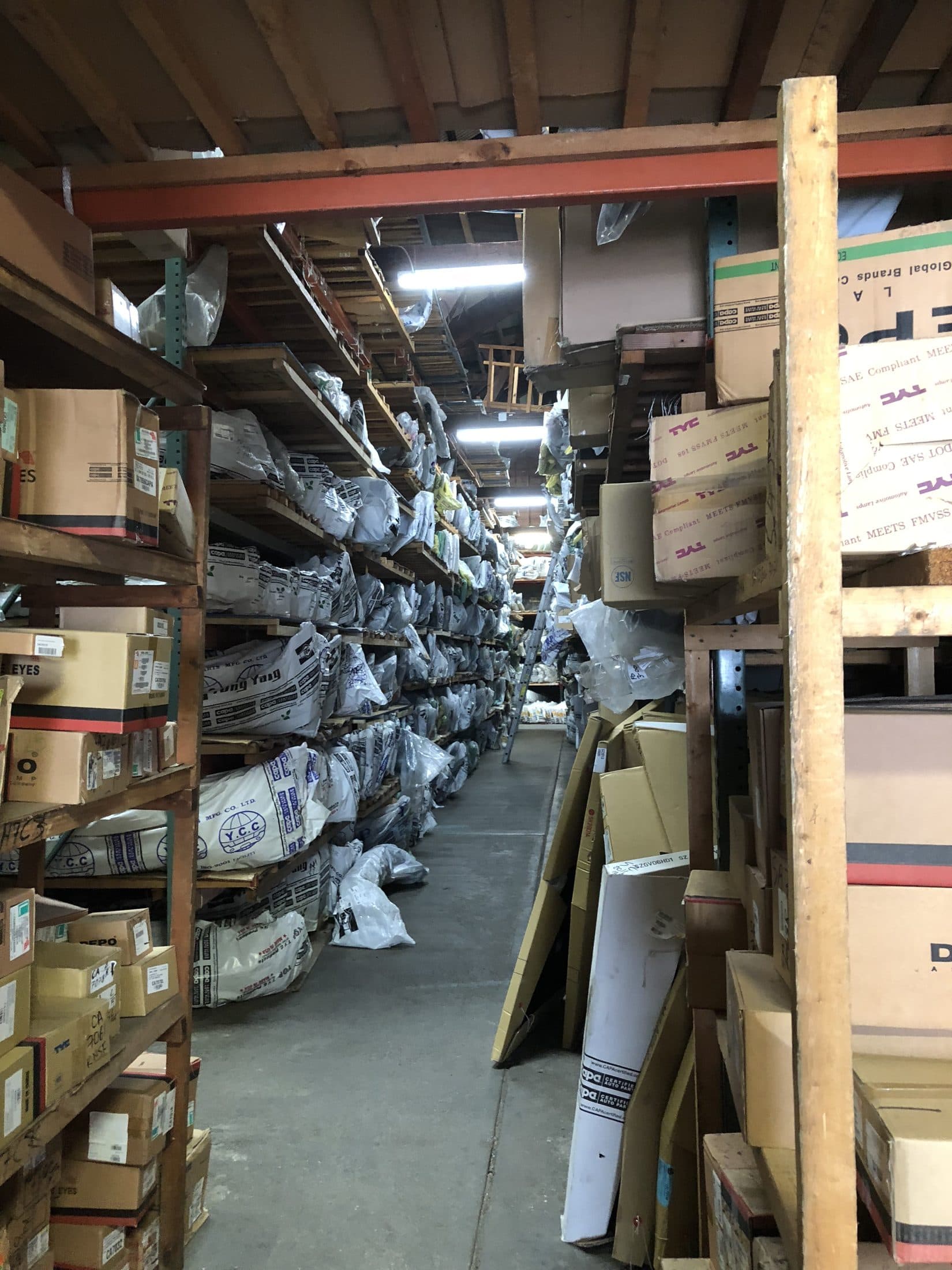 A narrow storeroom aisle with overfilled shelving and stock leaning into the walkway